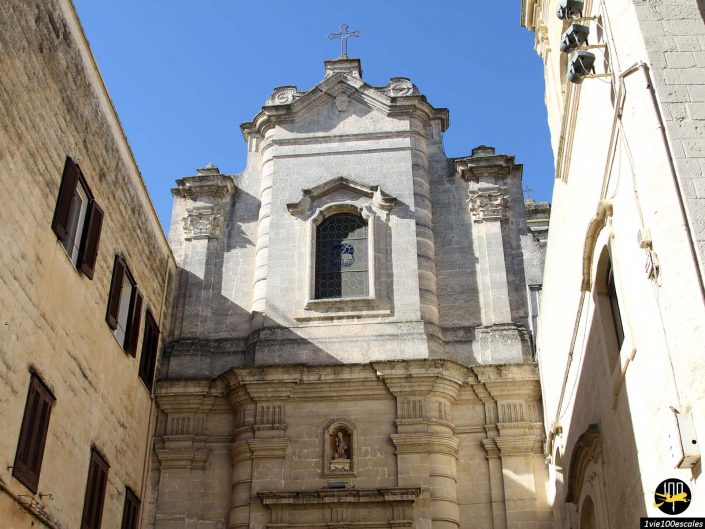 Vue de face d'une façade d'église en pierre avec des fenêtres cintrées, une petite statue dans une niche et une croix au sommet, située entre des bâtiments beiges sous un ciel bleu clair à Matera en Italie.