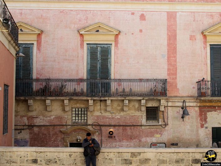 Une personne se tient seule sous un bâtiment rose patiné avec des volets et un balcon, regardant son téléphone. À Matera en Italie, la scène capture un moment de solitude. Un logo est visible dans le coin inférieur droit.