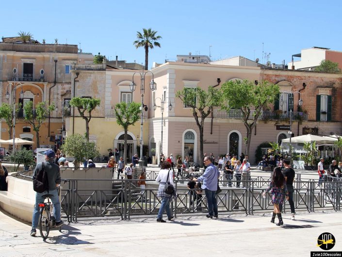 Les gens se rassemblent autour d'un espace extérieur avec des bâtiments historiques, des arbres et des lampadaires lors d'une journée ensoleillée à Matera en Italie. Certains marchent, d’autres sont assis, tandis que quelques-uns prennent des photos.