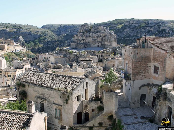 Des maisons anciennes en pierre et des structures aux toits de tuiles parsèment un paysage vallonné, probablement dans une vieille ville ou un quartier historique de Matera en Italie. Une formation rocheuse saisissante est visible en arrière-plan.