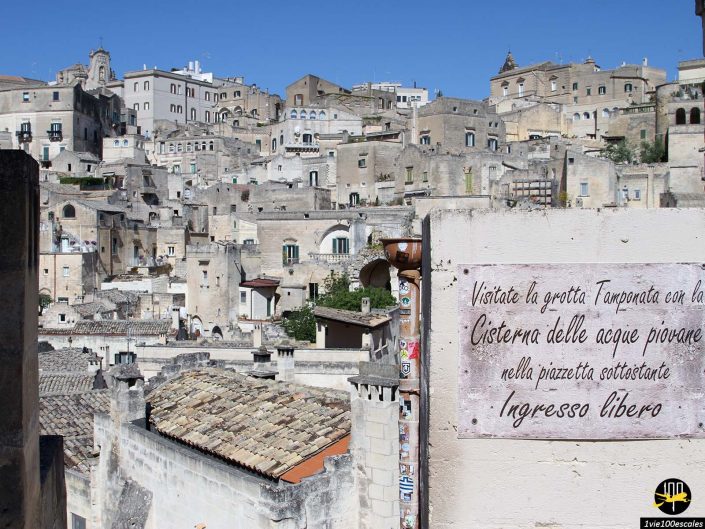 Un flanc de colline couvert de vieilles bâtisses en pierre à Matera en Italie, avec un panneau au premier plan indiquant "Visitez la grotte bien fermée avec la citerne d'eau de pluie sur la petite place devant. Entrée gratuite.