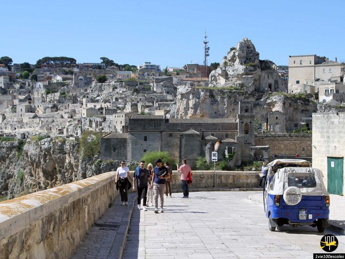 Les gens marchent le long d'un sentier en pierre avec des bâtiments historiques et des formations rocheuses en arrière-plan par temps clair à Matera en Italie. Un petit véhicule bleu est garé à droite.