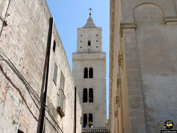 Vue d'un haut clocher en pierre de couleur claire avec trois niveaux de fenêtres cintrées, vu à travers l'étroit espace entre deux bâtiments en pierre plus anciens à Matera en Italie. Le ciel est clair et bleu.