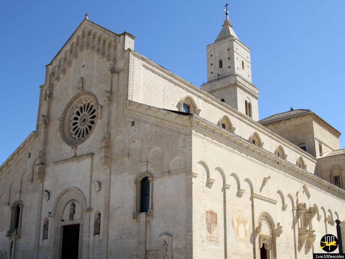 Une cathédrale historique en pierre beige avec des sculptures complexes et une grande rosace sous un ciel bleu clair se dresse fièrement à Matera en Italie. Une tour ornée s'élève élégamment au-dessus de la structure.