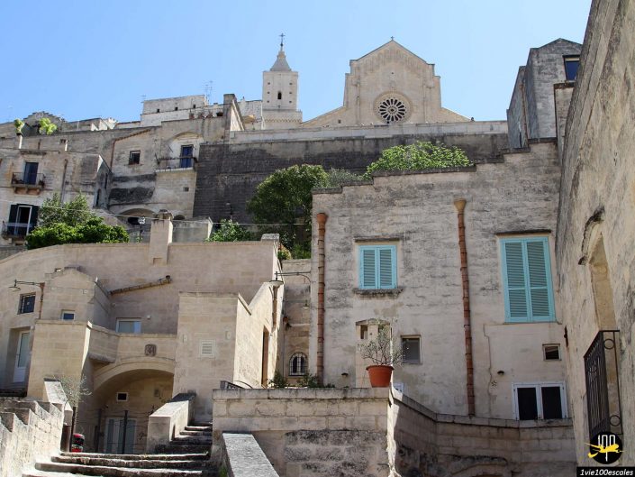Des bâtiments en pierre avec des volets pastel et des plantes en pot s'élèvent le long d'un chemin en escalier menant à une grande église avec une fenêtre circulaire proéminente sous le ciel bleu clair à Matera en Italie.