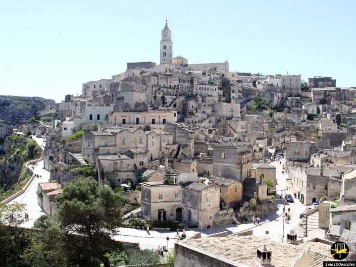 Une ville historique avec des bâtiments en pierre, des rues étroites et une haute tour centrale à flanc de colline sous un ciel clair à Matera en Italie.