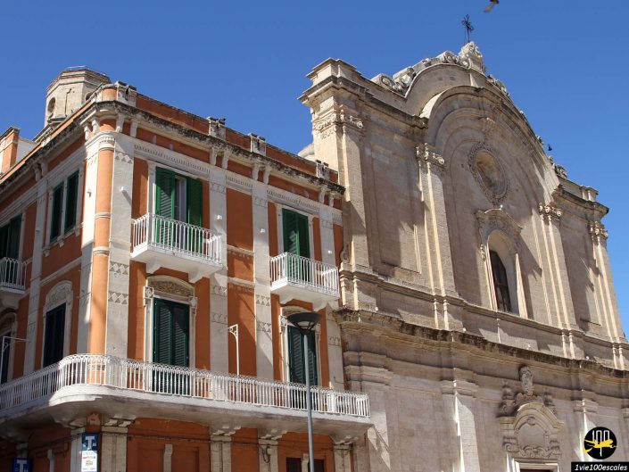 Un grand bâtiment en pierre orné de fenêtres cintrées et de détails complexes se dresse à côté d'un bâtiment orange adjacent aux volets verts et aux balcons blancs, sous un ciel bleu clair à Monopoli en Italie.