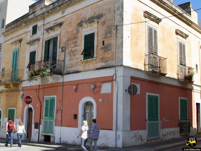 Bâtiment d'angle avec extérieur patiné, volets verts et petits balcons. Un panneau stop est visible sur le côté. Trois personnes passent devant l'immeuble dans la rue à Monopoli en Italie.