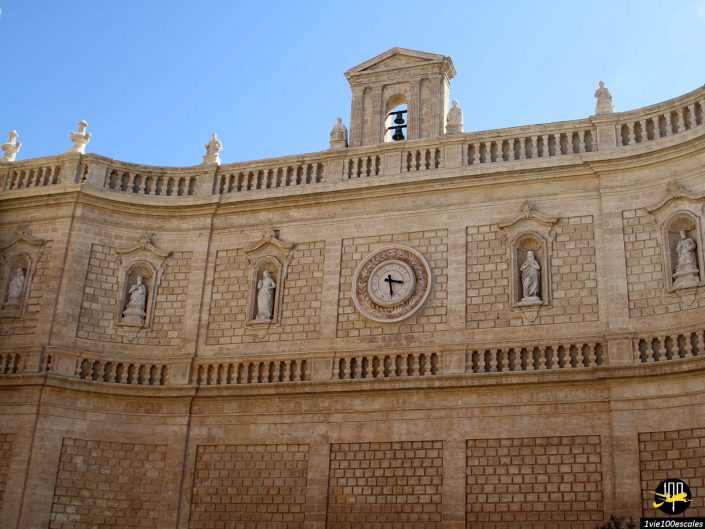 La façade d'un bâtiment en pierre à Monopoli, en Italie, présente des statues dans des alcôves, une horloge ronde au-dessus d'une croix et un petit beffroi avec une cloche au sommet, sur un ciel bleu clair.
