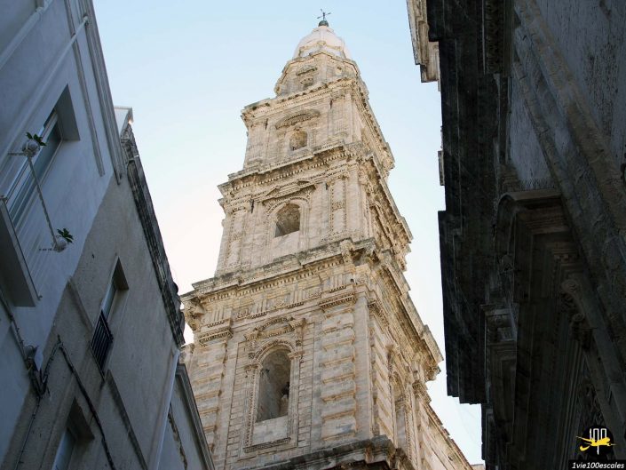 Un grand clocher en pierre orné s'élève entre deux bâtiments sous un ciel clair à Monopoli, en Italie, avec des fenêtres cintrées et des sculptures architecturales détaillées.