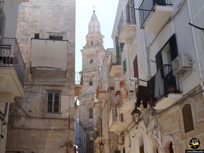 Une rue étroite bordée de bâtiments anciens mène à un haut clocher en pierre à Monopoli en Italie. Les vêtements sont suspendus aux balcons et une unité de climatisation est visible sur la droite. Le ciel est clair et ensoleillé.