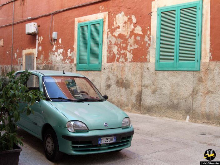 Une petite voiture verte est garée dans une rue calme à Monopoli en Italie, devant un immeuble orange patiné aux volets verts.