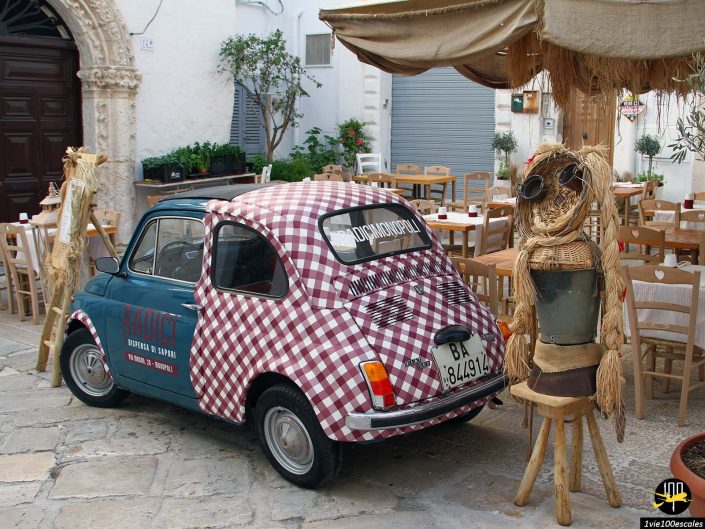 Une petite voiture vintage avec un motif à damier est garée près des tables à manger en plein air à Monopoli en Italie. Une silhouette ressemblant à un épouvantail se tient à côté de la voiture dans ce décor de rue pittoresque.