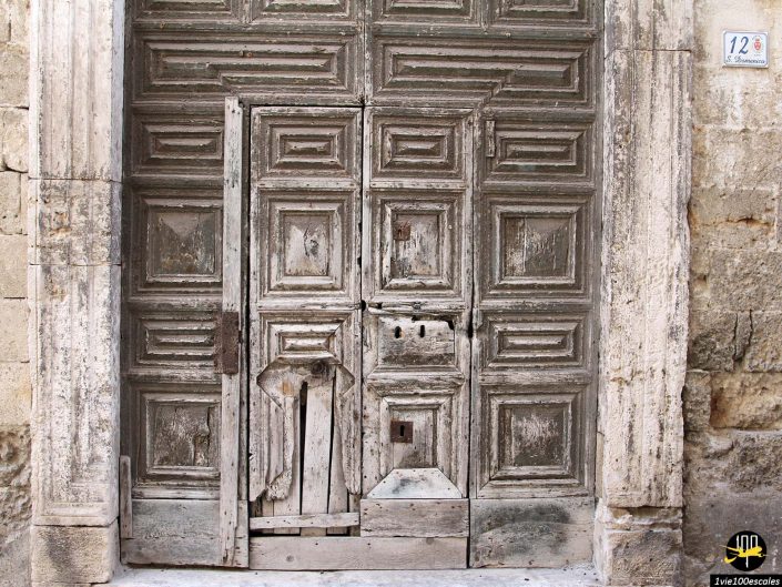 Une porte en bois patinée avec des panneaux géométriques complexes et des dommages visibles est montée dans un vieux mur de pierre à Monopoli en Italie. La porte comporte une petite partie inférieure barricadée et un panneau numéro 12 délavé au-dessus.