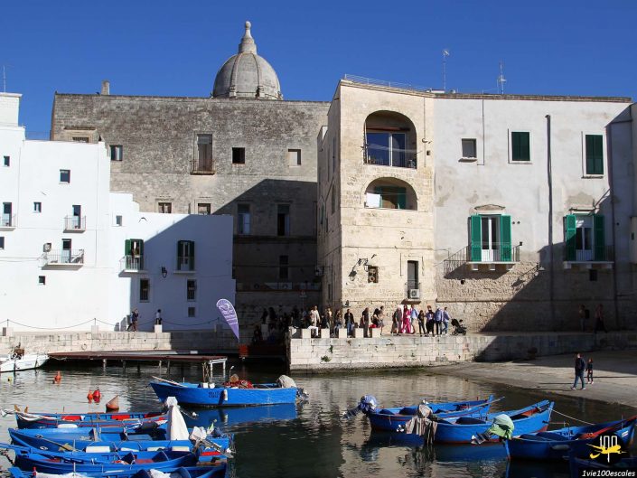 Un groupe de personnes se tient près d'un front de mer à Monopoli en Italie, devant des bâtiments historiques en pierre blanche et en pierre. Plusieurs bateaux bleus flottent sur l'eau au premier plan.