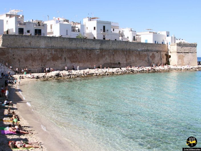 Une plage rocheuse aux eaux turquoise et claires côtoie un mur de pierre et des bâtiments blancs sous un ciel bleu clair à Monopoli en Italie. Les gens prennent le soleil, nagent et se détendent le long du rivage.