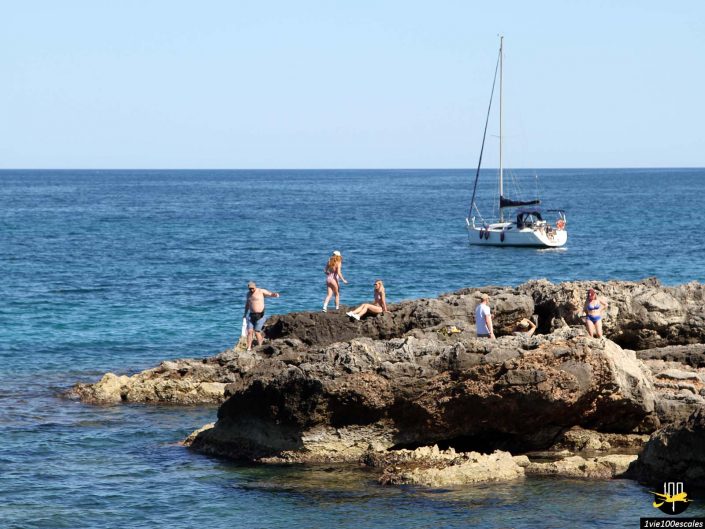 Les gens se détendent sur la côte rocheuse près de l'océan à Monopoli en Italie, avec un voilier ancré en arrière-plan par une journée ensoleillée.