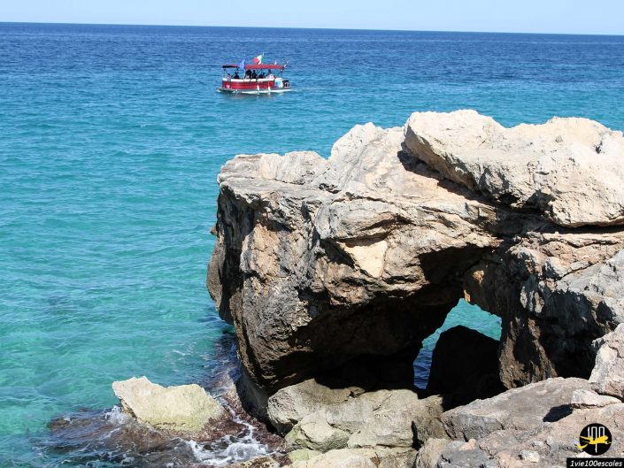 Un petit bateau flotte sur une eau bleu clair près d'un rivage rocheux avec de grandes formations rocheuses à Monopoli en Italie.