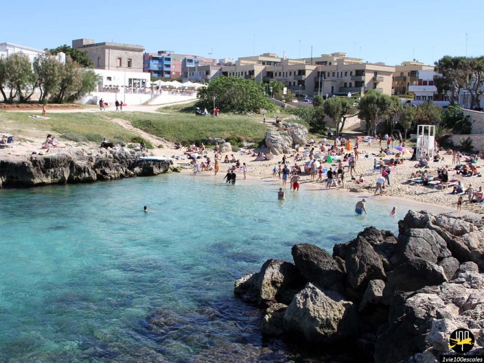 Une scène de plage à Monopoli en Italie avec des gens bronzant, nageant et se relaxant près d'une eau turquoise claire. Des bâtiments et des arbres sont visibles en arrière-plan. Des rochers et une tour de secours sont présents sur le rivage.