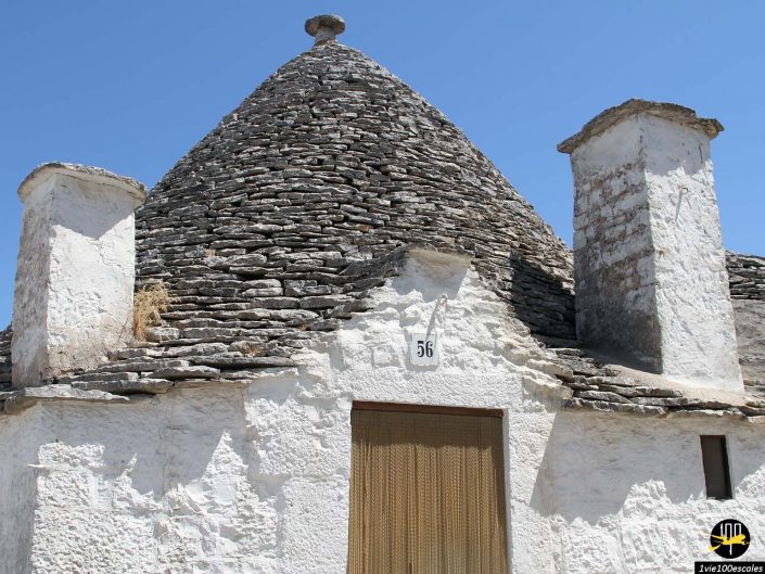 Une maison trullo traditionnelle avec un toit conique en pierre et des murs blanchis à la chaux, avec le numéro 56 au-dessus de la porte, sous un ciel bleu clair à Alberobello en Italie.