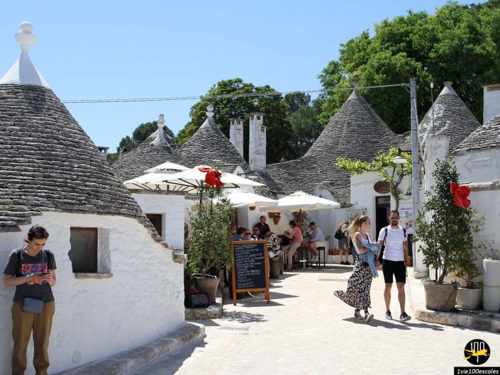 Des gens marchant et assis dans un café extérieur ensoleillé entouré de bâtiments trullo blancs traditionnels aux toits coniques à Alberobello en Italie.