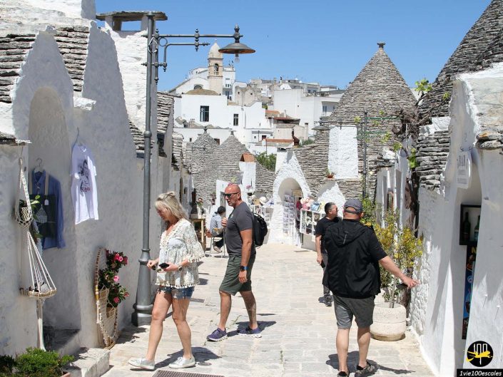 Les gens marchent dans une rue étroite bordée de maisons trulli traditionnelles blanchies à la chaux et aux toits coniques à Alberobello en Italie. Les vendeurs ambulants exposent des objets et le ciel est clair et bleu.
