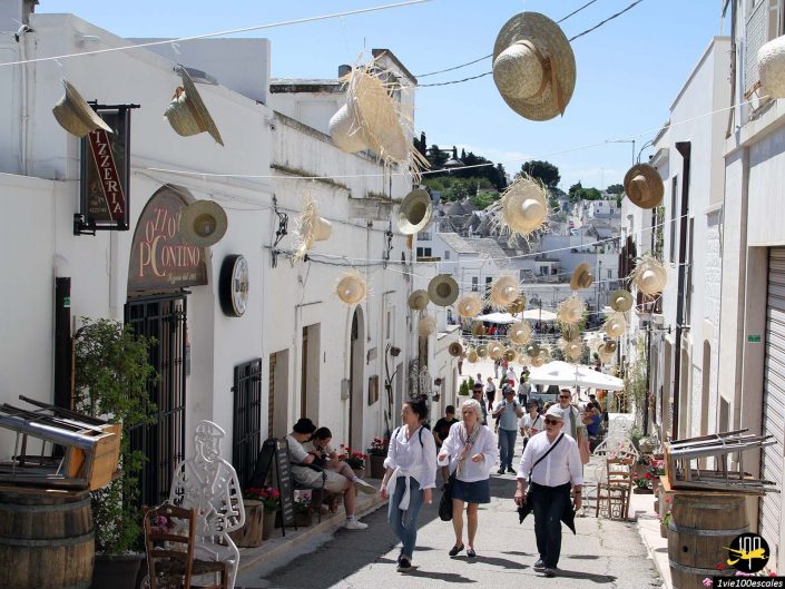 Une rue étroite à Alberobello en Italie est décorée de chapeaux de paille suspendus. Les gens marchent et s’assoient devant les cafés, tandis que des bâtiments blancs bordent la rue. Une charmante enseigne de restaurant est visible sur la gauche.