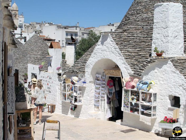 Une rue étroite bordée de maisons Trulli blanches aux toits coniques, exposant divers souvenirs et objets artisanaux. Une personne portant un chapeau et un short marche le long du chemin sous la lumière du soleil, à Alberobello en Italie.