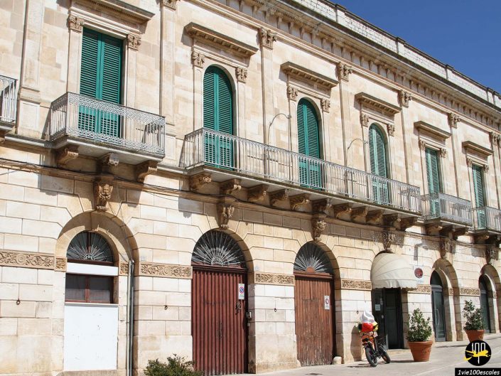 Un bâtiment historique avec des volets verts et deux grandes portes en bois au rez-de-chaussée, une moto garée à proximité et plusieurs plantes en pot devant à Locorotondo en Italie.