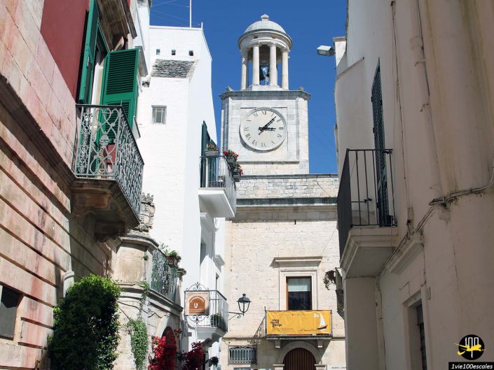 Une petite ruelle dans une ville historique à Locorotondo en Italie avec une tour d'horloge avec un grand cadran d'horloge. Des immeubles avec balcons et volets verts bordent la rue étroite sous un ciel bleu clair.