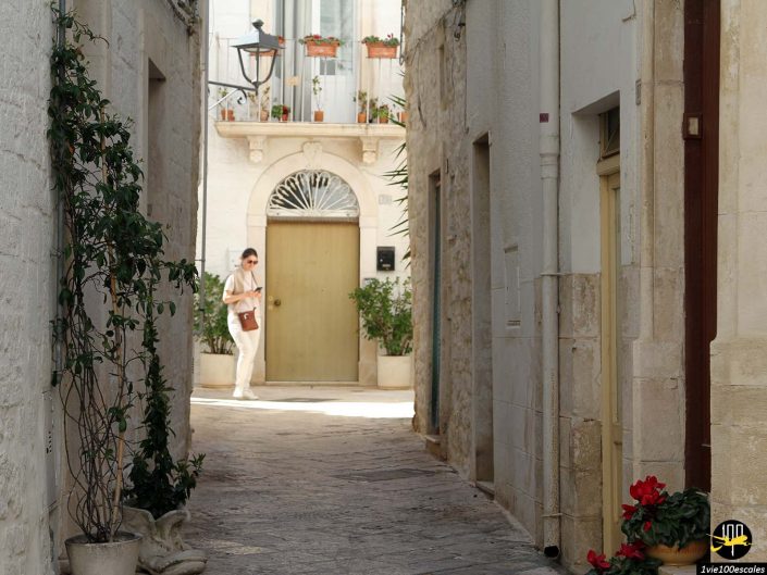 Une ruelle étroite et ensoleillée avec des plantes en pot mène à une cour où une personne en tenue blanche avec des lunettes de soleil et un sac marron se dirige vers une porte jaune, à Locorotondo en Italie.