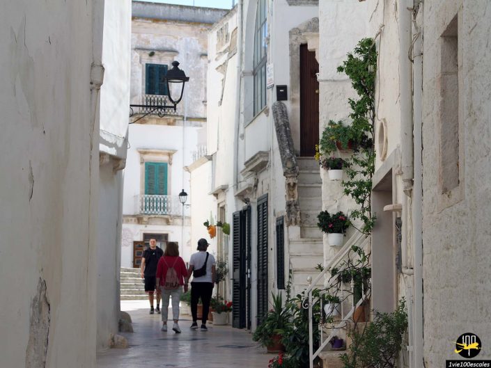 Une rue pavée étroite entre des bâtiments blancs avec des plantes suspendues, trois personnes marchant et un lampadaire orné à Locorotondo en Italie.