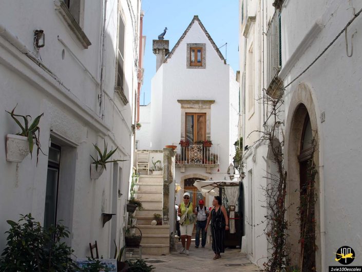 Rue piétonne étroite dans une ville blanchie à la chaux à Locorotondo en Italie avec des bâtiments en pierre, des plantes en pot et des escaliers ; deux personnes marchent au centre, l’une tenant un appareil photo. Le ciel est clair et bleu.