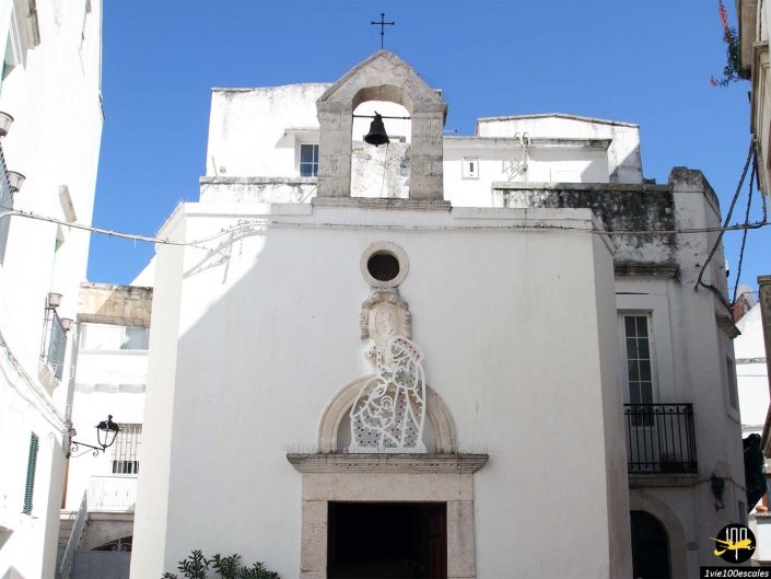 Une petite chapelle blanche avec un clocher, une fenêtre circulaire et un relief complexe en pierre sculptée au-dessus de l'entrée se dresse à Locorotondo en Italie. Le bâtiment est entouré de structures blanches sous un ciel bleu clair.
