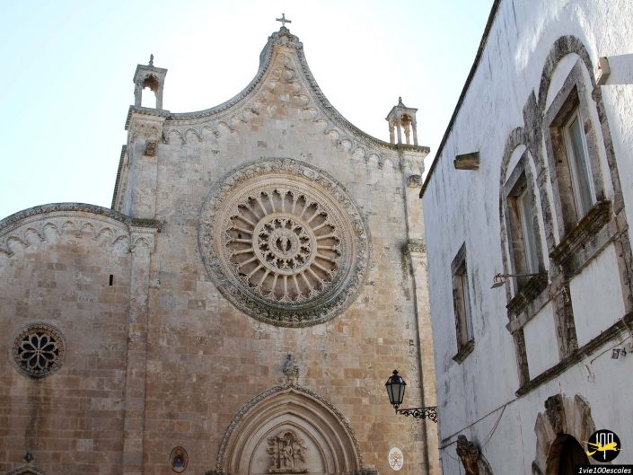 Façade de cathédrale historique en pierre avec une grande rosace et des sculptures détaillées, adjacente à un bâtiment blanc à Ostuni en Italie. Un lampadaire est installé sur le bâtiment blanc, avec un ciel bleu clair en arrière-plan.