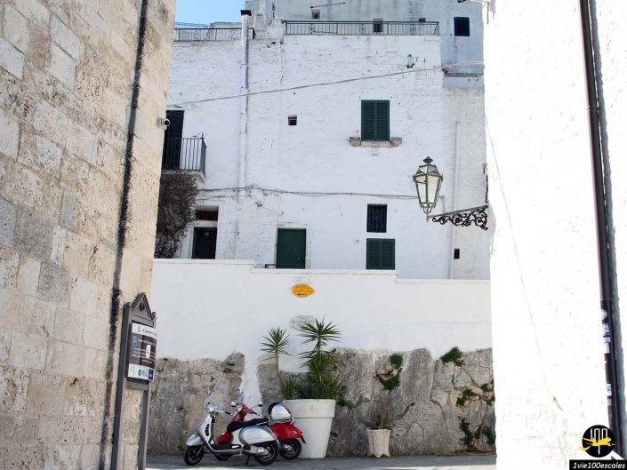 Deux scooters sont garés près d’un mur de pierre blanche avec une plante en pot. Il y a un bâtiment en pierre sur la gauche et des bâtiments blancs à plusieurs niveaux en arrière-plan. Un lampadaire est fixé au mur, capturant l'essence de la vie quotidienne à Ostuni en Italie.