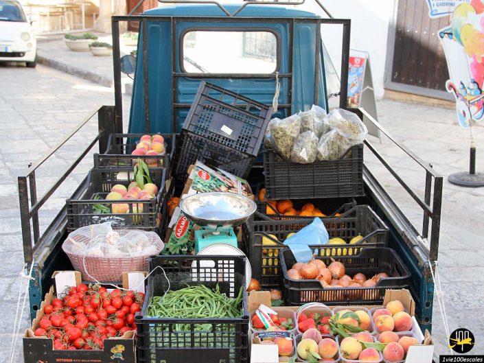 Un petit camion transporte des caisses remplies de divers fruits et légumes frais, notamment des tomates, des pêches, des haricots verts et un panier de légumes verts dans une rue de la ville d'Ostuni en Italie.