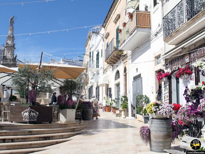 Une scène de rue ensoleillée dans une vieille ville européenne à Ostuni en Italie avec des terrasses de cafés, des plantes en pot, des fleurs colorées et des bâtiments historiques. Un grand monument est visible en arrière-plan sous un ciel bleu clair.