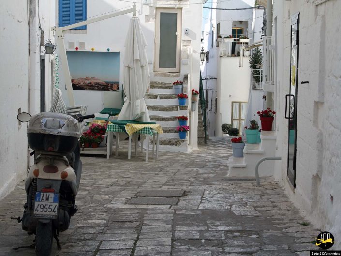 Une rue pavée étroite dans un village méditerranéen à Ostuni en Italie avec des bâtiments blancs, des pots de fleurs colorés, un scooter garé et des tables avec parasols et chaises installées sous un escalier.