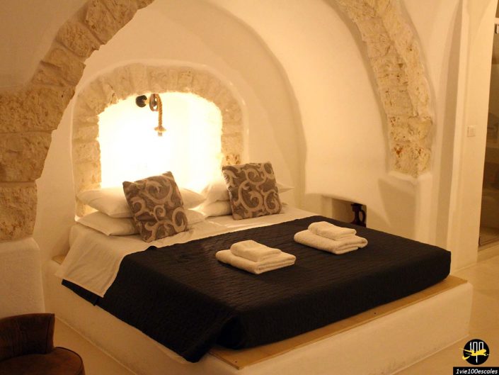 Une chambre cosy à Ostuni en Italie avec une arche en pierre au dessus du lit, éclairée par une petite applique murale. Le lit est fait de draps blancs, de deux oreillers à motifs, d'un couvre-lit sombre et de serviettes soigneusement pliées au pied.