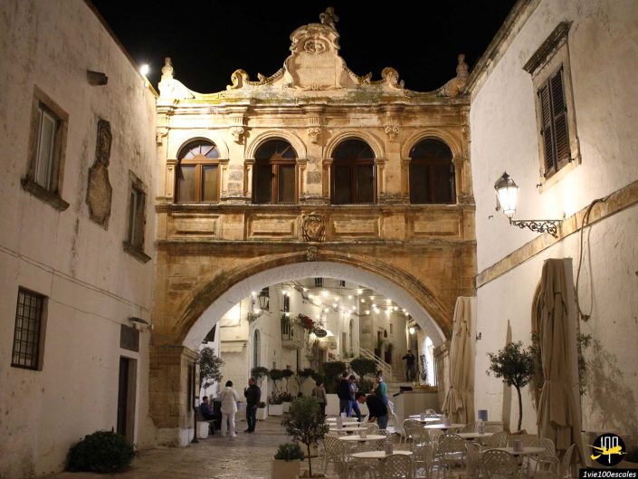 Une arche historique avec des pierres architecturales détaillées s'étend sur une rue étroite bordée de bâtiments blancs et de sièges extérieurs, éclairée la nuit à Ostuni en Italie. Les gens sont rassemblés sous la voûte.