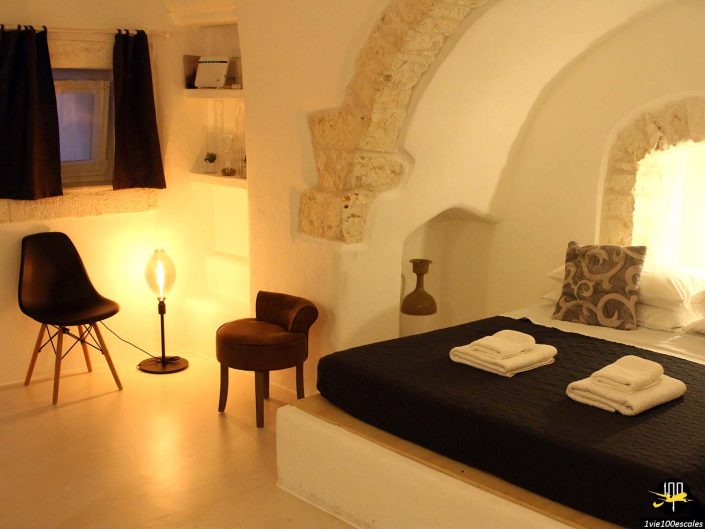 Une chambre confortable à Ostuni en Italie avec des arches en pierre et un éclairage chaleureux. Il comprend un lit avec une literie sombre, deux serviettes pliées, une petite chaise, une chaise noire, une lampe de table d'appoint et des étagères avec des objets décoratifs.