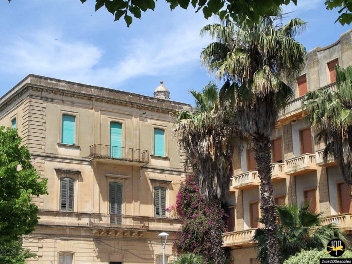 Deux bâtiments beiges à plusieurs étages aux volets turquoise se dressent à Lecce en Italie, entourés d'une verdure luxuriante et de palmiers. Le ciel est clair et bleu.