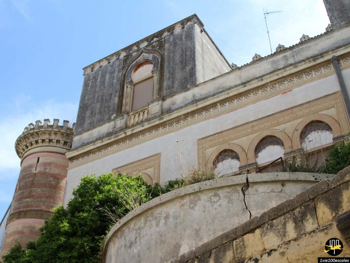 Un château en pierre partiellement détérioré avec une tour cylindrique et des fenêtres cintrées, partiellement obscurcies par la verdure, se dresse majestueusement à Lecce en Italie. Ciel bleu vif en arrière-plan.