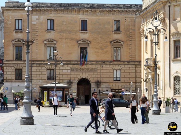 Une petite foule est rassemblée sur une place ensoleillée de Lecce en Italie avec des bâtiments historiques en arrière-plan, dont le « Palazzo Di Giustizia ». Des piétons marchent et deux personnes conversent au premier plan.