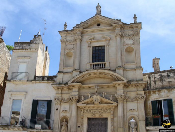 Un bâtiment historique en pierre beige avec des détails architecturaux ornés et des sculptures au-dessus de l'entrée, sur un ciel bleu clair, à Lecce en Italie.