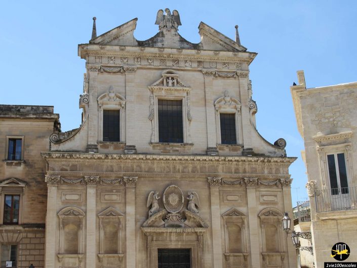 Un bâtiment historique en pierre avec des détails architecturaux ornés, notamment un fronton triangulaire avec une sculpture d'aigle, de grandes fenêtres cintrées et des sculptures décoratives autour de l'entrée et des fenêtres, se dresse fièrement à Lecce en Italie.