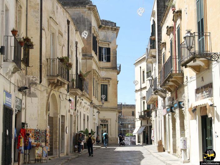 Une rue étroite d'une vieille ville européenne, à Lecce en Italie, bordée de bâtiments historiques, de balcons et de quelques personnes marchant ou assises dans un café. Des panneaux et des décorations sont visibles, avec un ciel clair au-dessus.