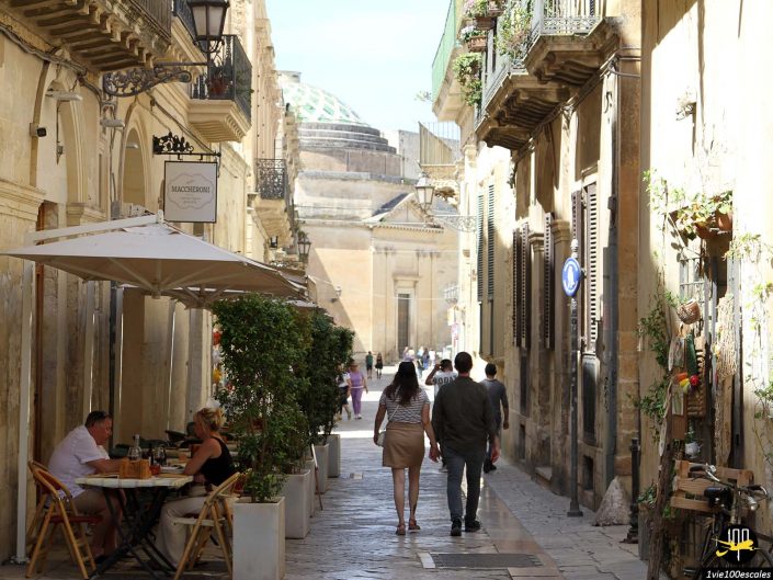 Une rue étroite et ensoleillée de Lecce en Italie est bordée de vieux bâtiments et de plantes. Les gens marchent et s’assoient à des tables extérieures. Un grand dôme est visible en arrière-plan.