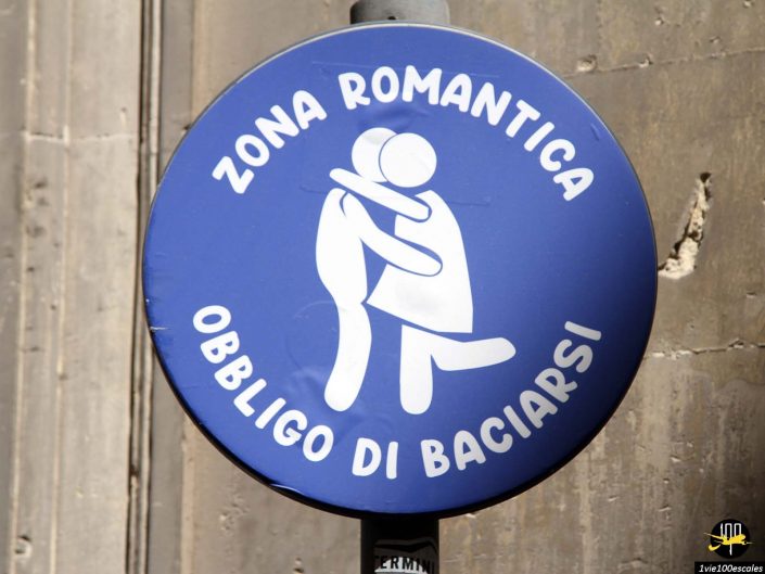 Un panneau circulaire bleu avec deux personnages en bâton blanc s'embrassant. Le panneau indique « Zona Romantica Obbligo di Baciarsi », ce qui se traduit de l'italien par « Zone romantique obligation de s'embrasser », à Lecce en Italie.